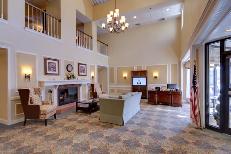 A spacious and well-lit living room area with high ceilings, a chandelier, patterned carpet, a fireplace with framed pictures and a clock above it, two upholstered armchairs, a sofa, a coffee table, a wooden desk with a computer, and an American flag near the glass entrance door.