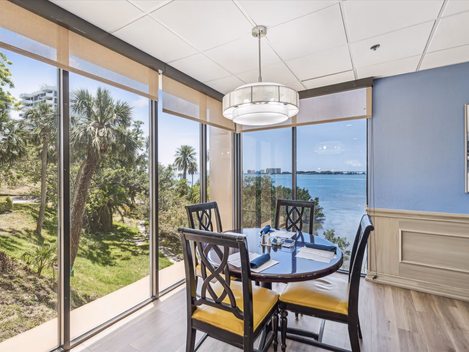 A dining area with a round table set for four, featuring dark wooden chairs with yellow cushions. The room has large floor-to-ceiling windows offering a scenic view of palm trees, greenery, and a body of water under a clear blue sky. A modern circular light fixture hangs above the table, and the walls are painted blue with beige wainscoting.