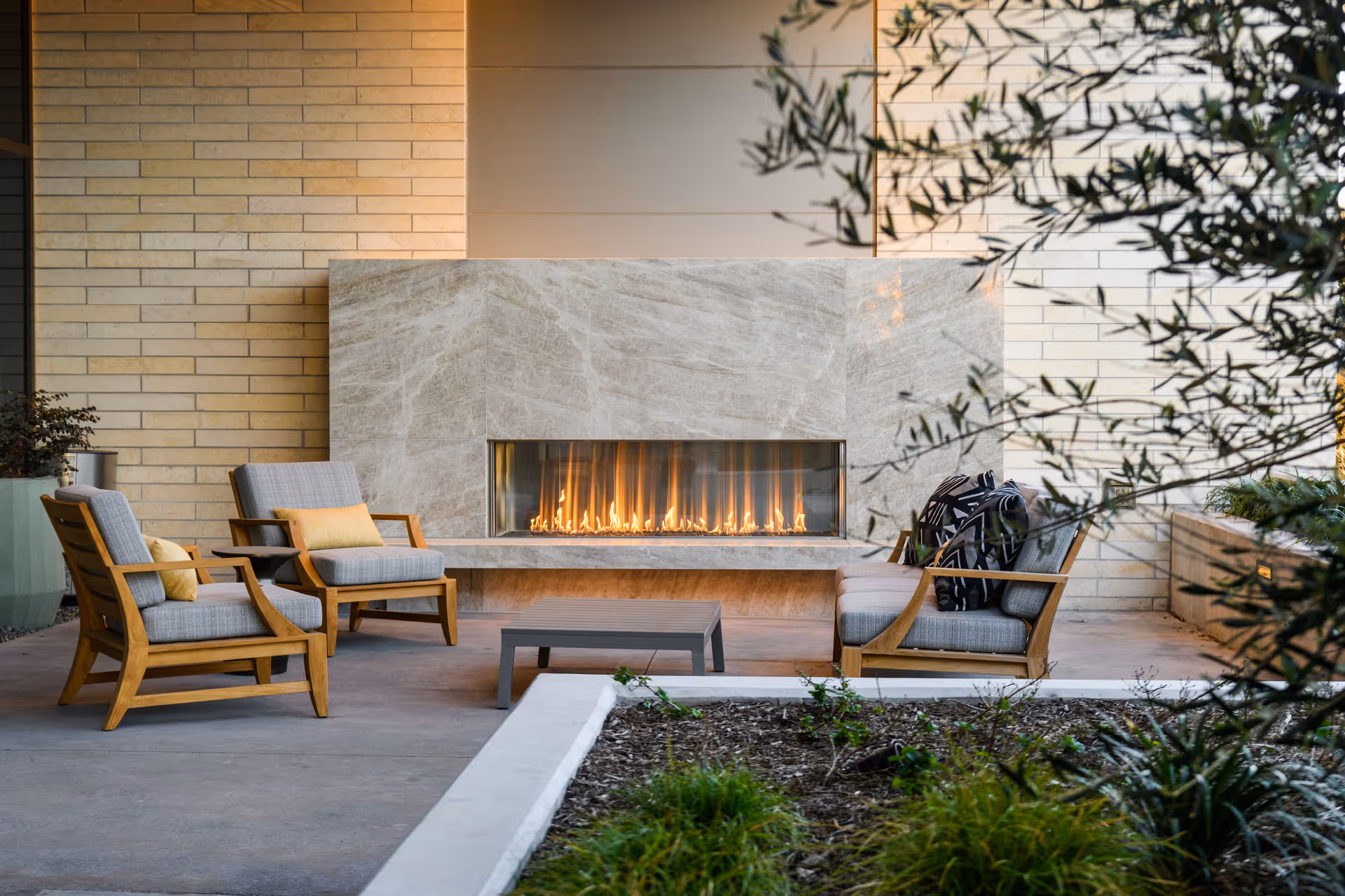 Outdoor seating area with four cushioned wooden chairs arranged around a modern rectangular fireplace built into a stone wall. There is a small coffee table in the center and some greenery in the foreground and background.