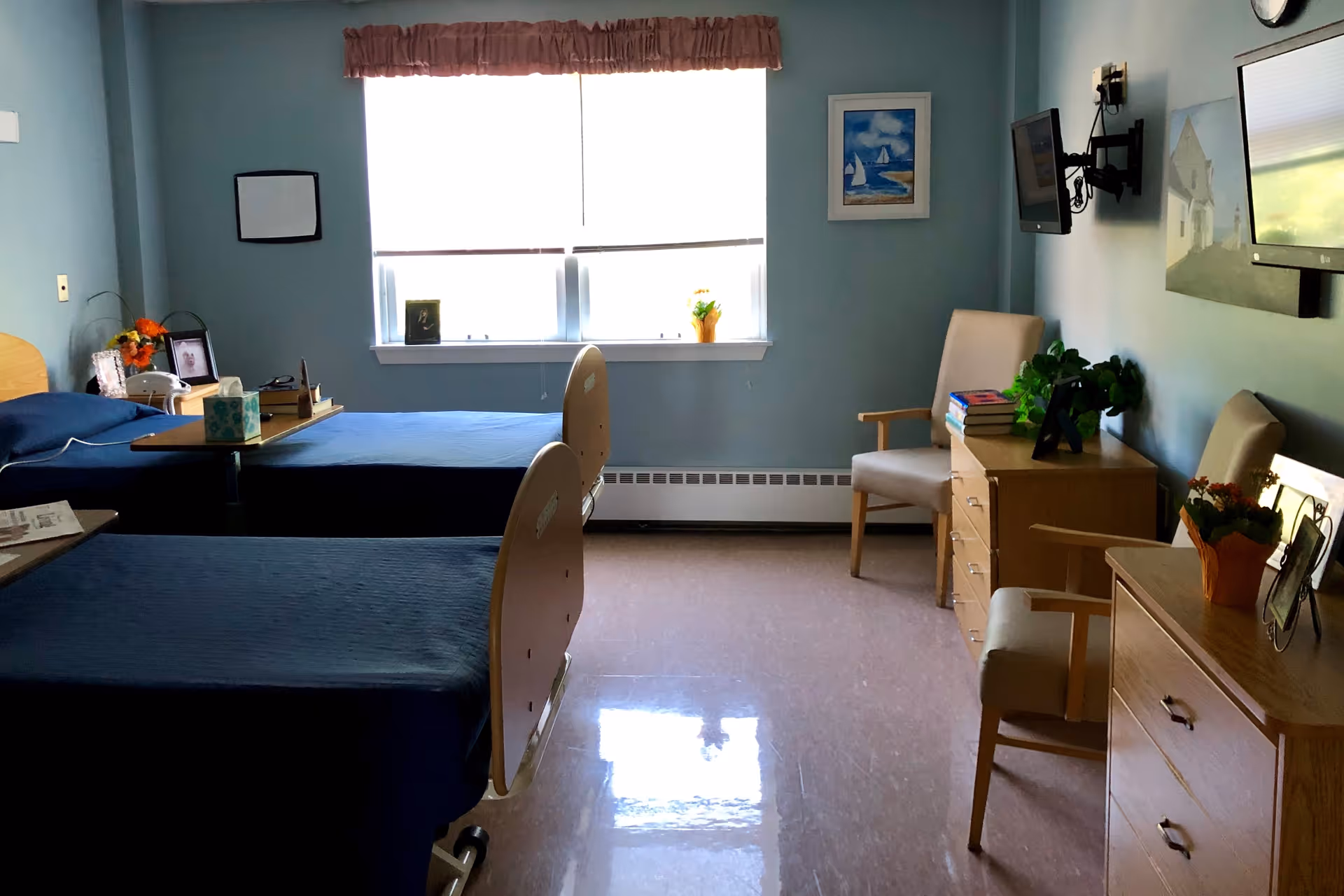 A shared bedroom in a healthcare facility with two hospital-style beds covered in blue bedding. The room has light blue walls, a window with a valance, two chairs, two dressers with plants and books on top, and two wall-mounted televisions. There are framed pictures on the walls and on the dressers.