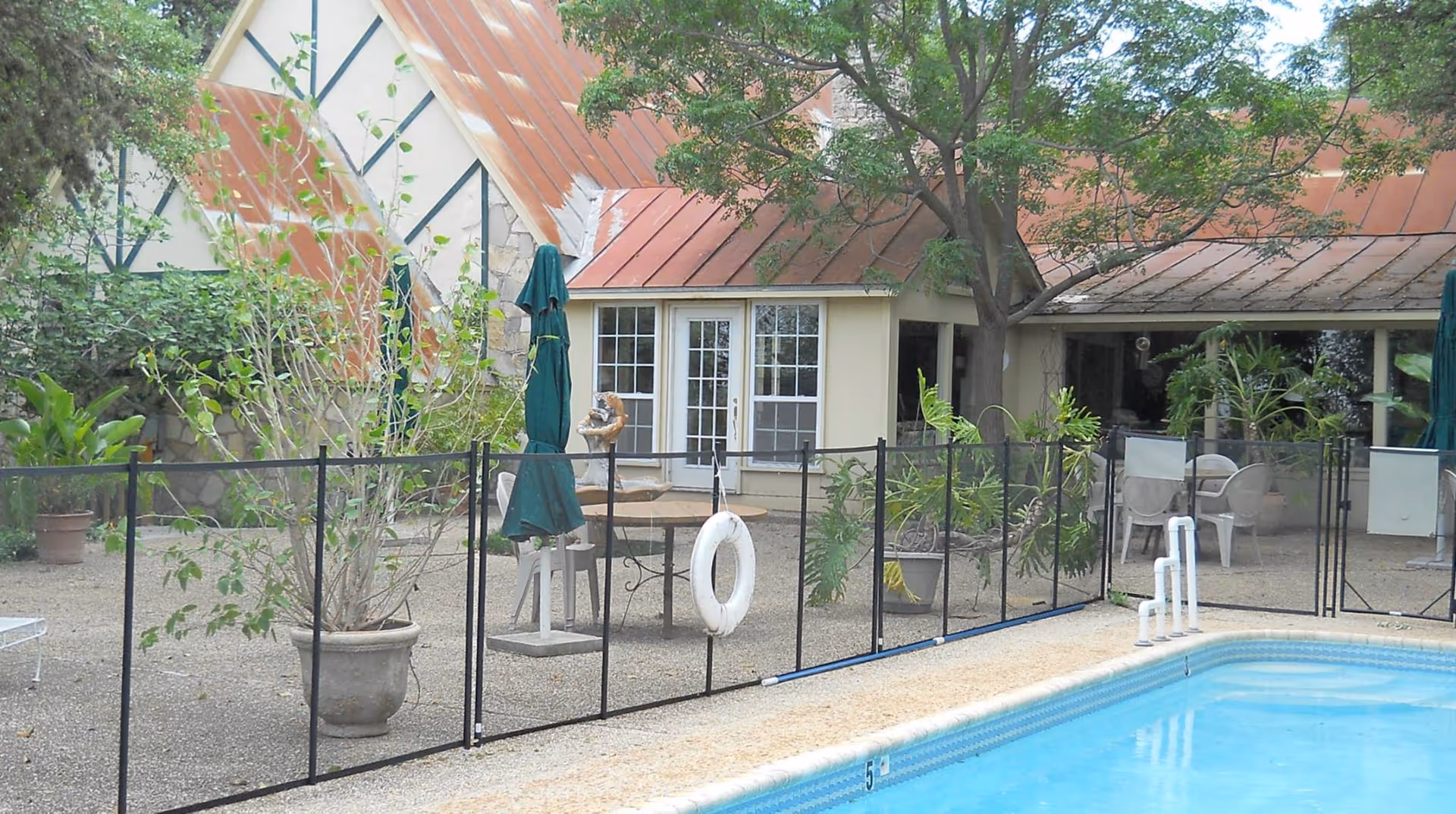 Outdoor pool area with a safety fence, patio furniture, potted plants, and a building with a rust-colored metal roof in the background.