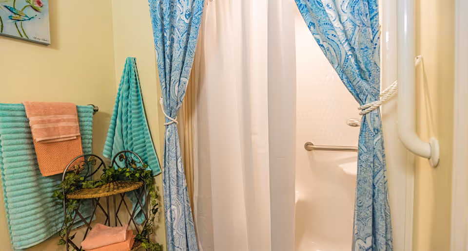 A bathroom shower stall with blue patterned curtains and a white liner, a grab bar inside, and folded teal and peach towels on a nearby rack with decorative greenery.