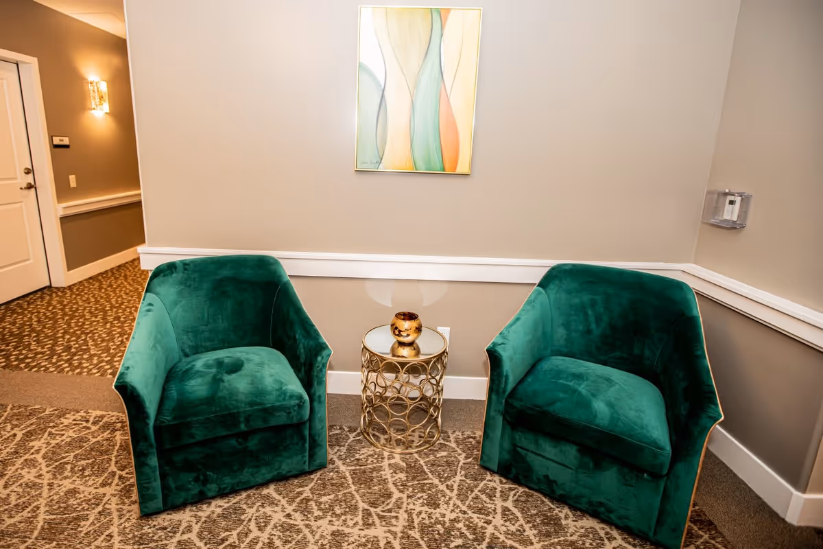 Two green velvet armchairs with a small round gold side table between them, topped with a decorative item, set against a beige wall with abstract artwork hanging above. The setting appears to be a hallway or common area with patterned carpet and a door visible in the background.
