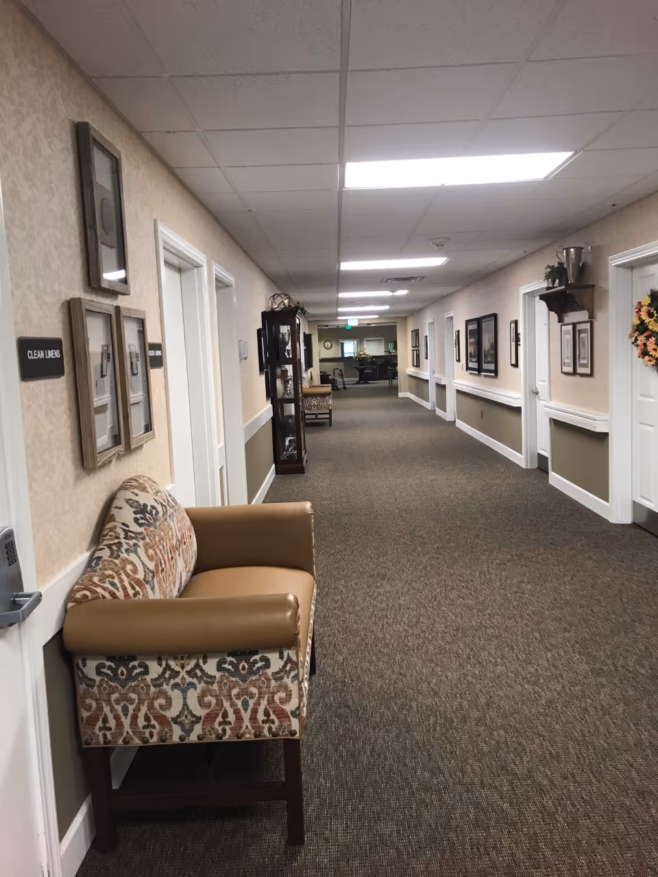 A long, carpeted hallway in an assisted living facility with beige walls and white trim. There are several closed white doors along the hallway, framed pictures and decorations on the walls, and a patterned upholstered bench with tan armrests near the foreground. The ceiling has fluorescent lights, and there is a cabinet with decorative items further down the hall.
