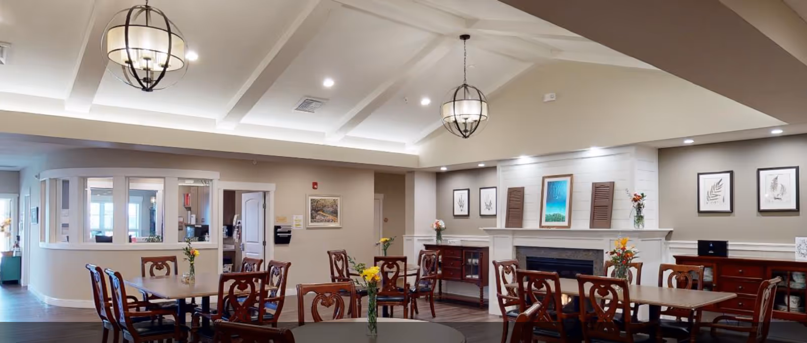 Communal dining room with several wooden tables and chairs, a fireplace with artwork, and hanging pendant lights.