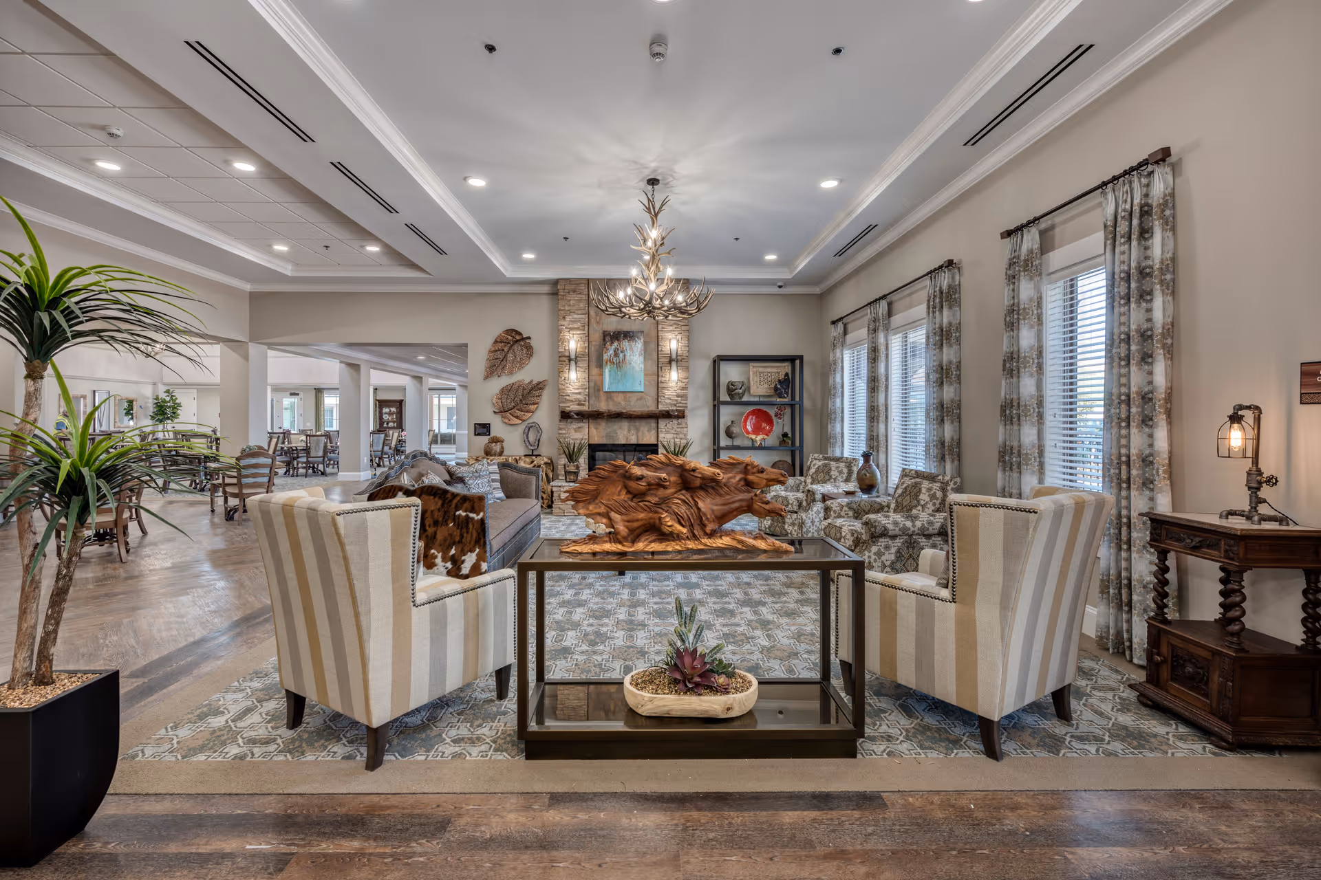 A spacious and elegantly decorated senior living facility common area with comfortable striped armchairs, a wooden sculpture centerpiece on a glass table, patterned curtains on large windows, a stone fireplace with wall art above it, and additional seating in the background. The room features warm lighting and a mix of modern and rustic decor elements.