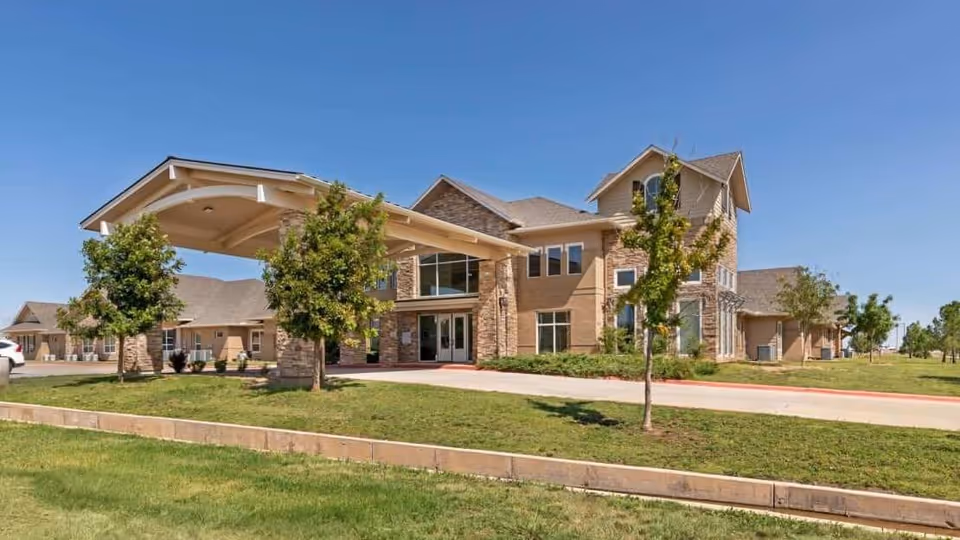Exterior view of Bristol Park at Amarillo Assisted & Living Memory Care facility showing a large, two-story building with stone and beige siding, a covered entrance, several windows, small trees, and a well-maintained lawn under a clear blue sky.