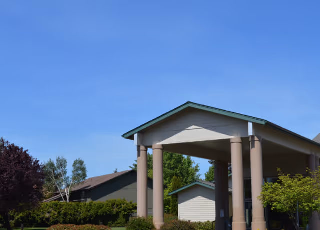 Entrance of a building with a covered portico supported by four large columns, surrounded by green trees and bushes under a clear blue sky.