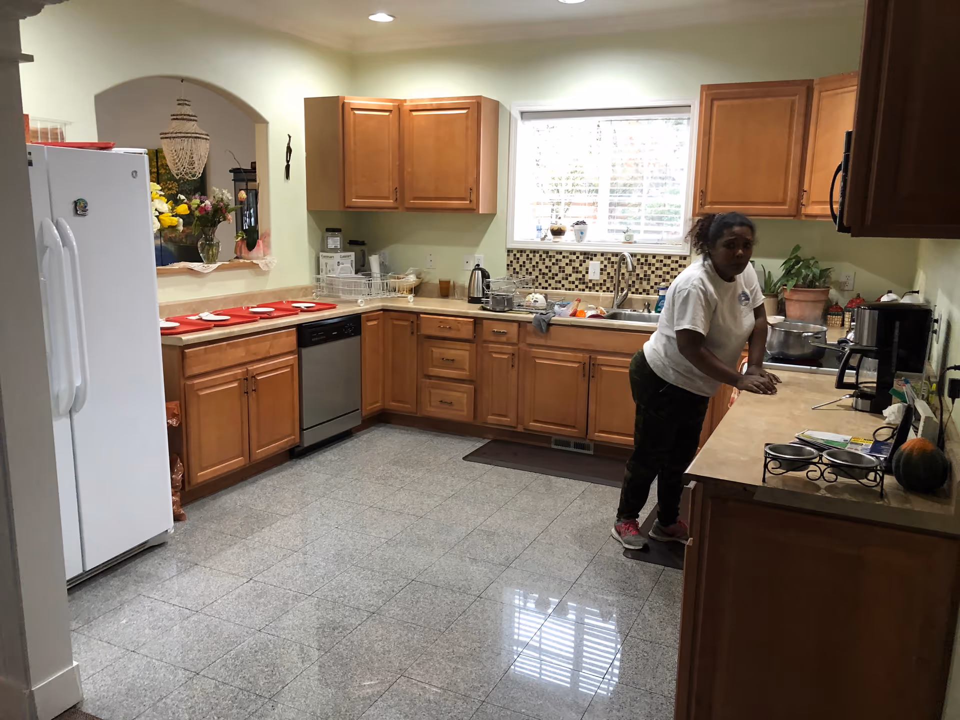 A woman standing in a kitchen with wooden cabinets, a white refrigerator, a dishwasher, and a window above the sink. The kitchen has a tiled floor and various kitchen items on the countertops.