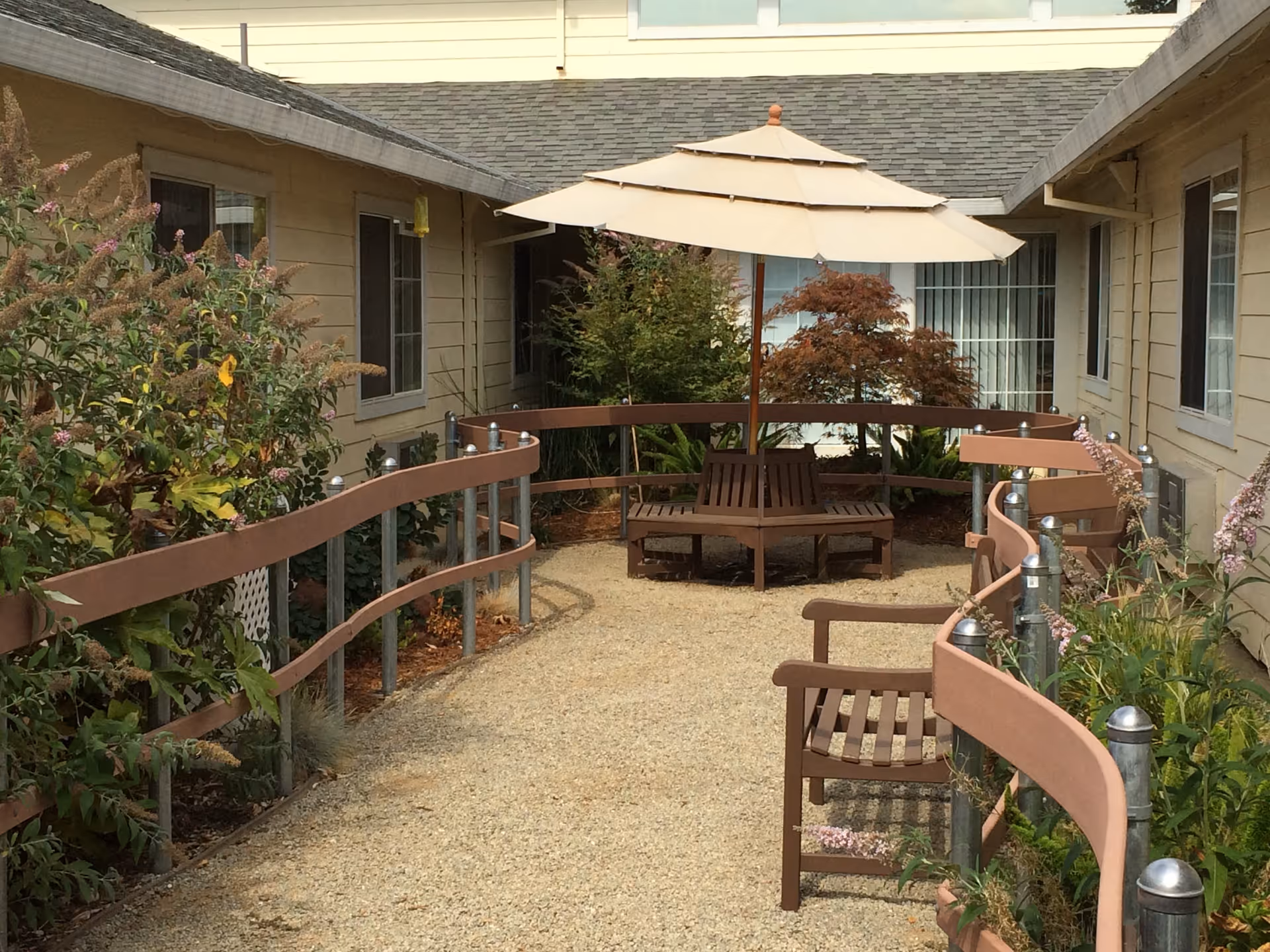 Outdoor courtyard area with a gravel pathway, brown wooden benches, a round wooden bench with a beige umbrella in the center, surrounded by plants and shrubs, and beige building walls with windows on both sides.
