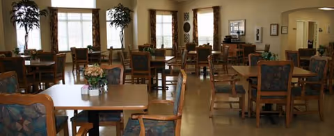 Spacious dining room with multiple wooden tables and upholstered chairs, large windows, and potted plants.