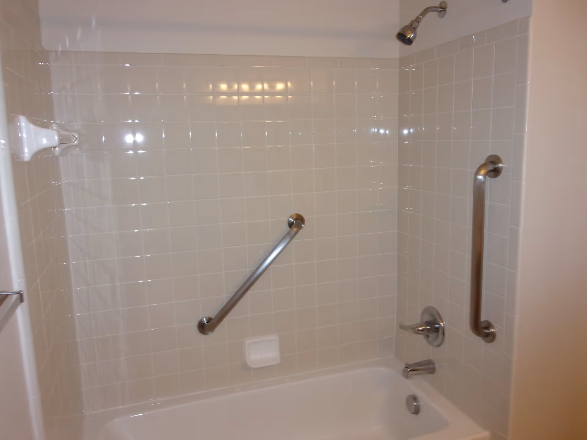 A clean bathroom with a white bathtub and tiled walls. The bathtub has two metal grab bars, one horizontal and one vertical, for safety. There is a showerhead mounted on the wall above the tub and a soap holder built into the tiled wall.