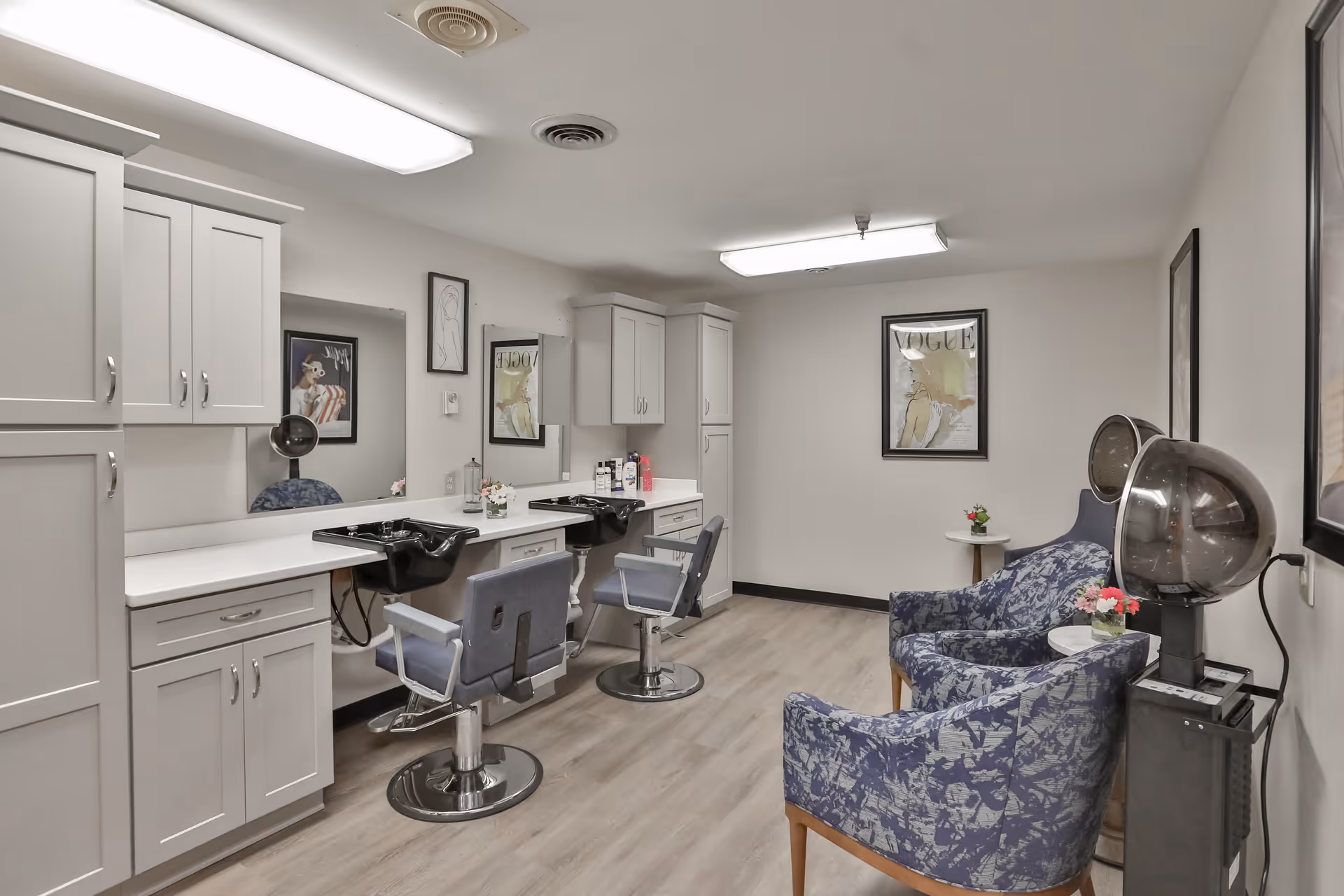 Interior view of a senior living facility's hair salon area featuring two salon chairs with sinks, white cabinetry, a large mirror, and two blue patterned armchairs with a small table between them. The room has light wood flooring, framed artwork on the walls, and a hair dryer machine.