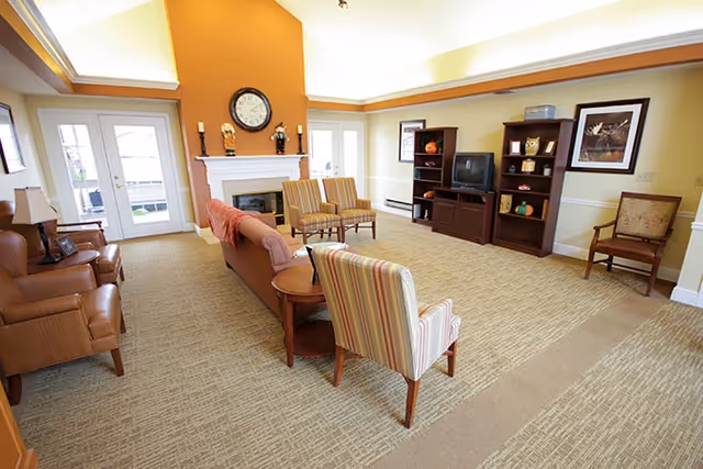 A spacious living room with beige carpet and light yellow walls. The room features a white fireplace with a clock and decorative items on the mantel, two striped armchairs, a brown sofa with a pink throw, two leather armchairs, a wooden side table, and a wooden entertainment center with a TV and decorative items. French doors with glass panes lead outside, and framed artwork hangs on the walls.