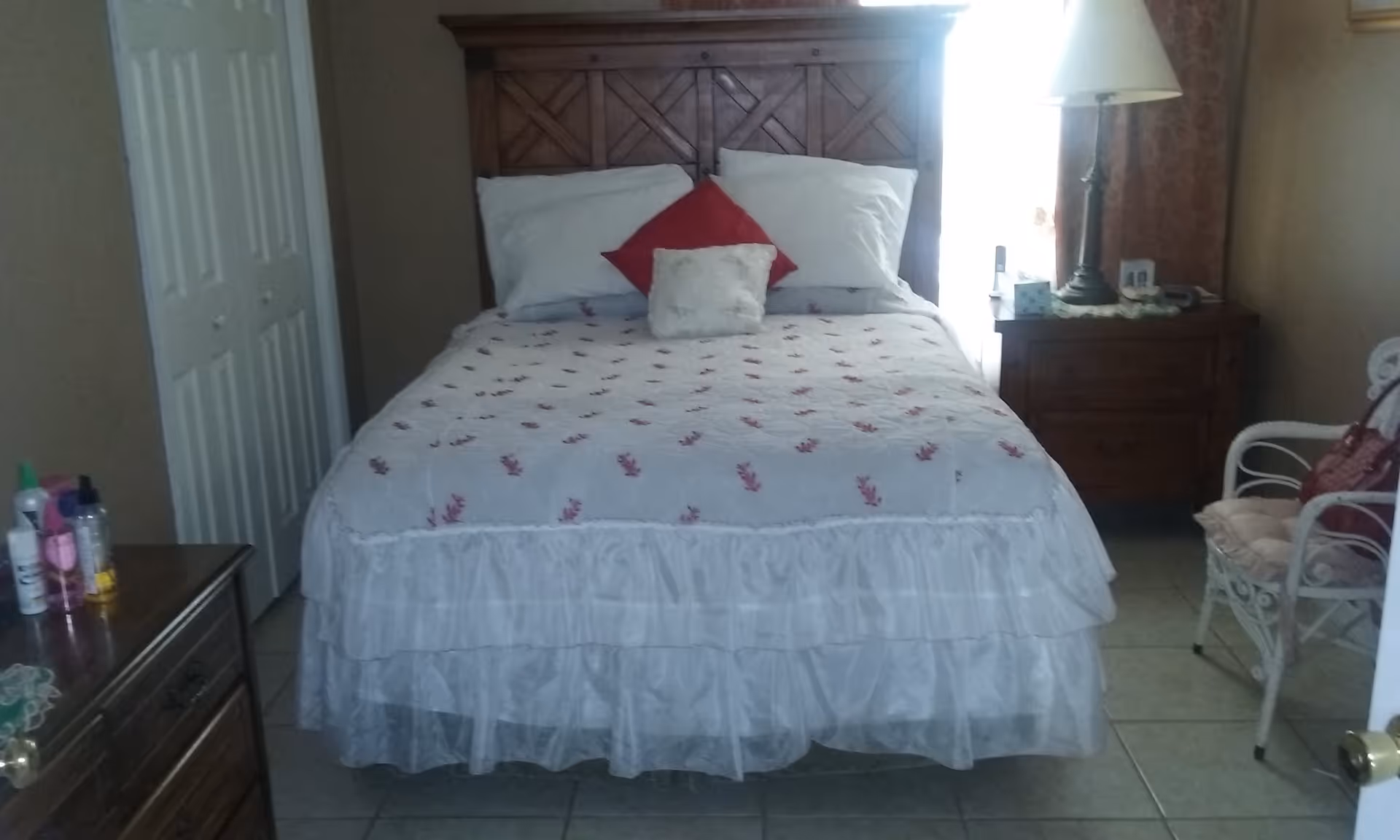 Neatly made bed with a wooden headboard, pillows and a ruffled bedskirt in a modest bedroom with a nightstand, lamp and dresser.