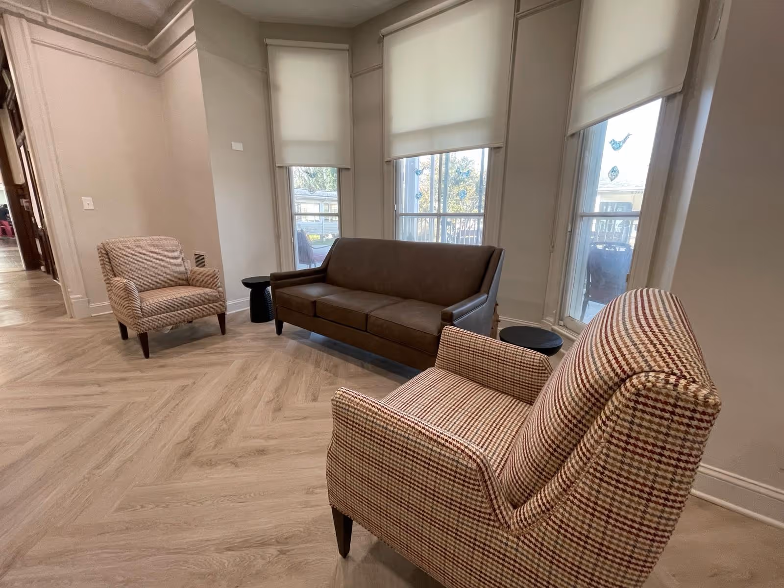 A sitting area with a brown leather sofa and two patterned armchairs arranged around two small black side tables. The room has large windows with white roller shades and light wood herringbone flooring.