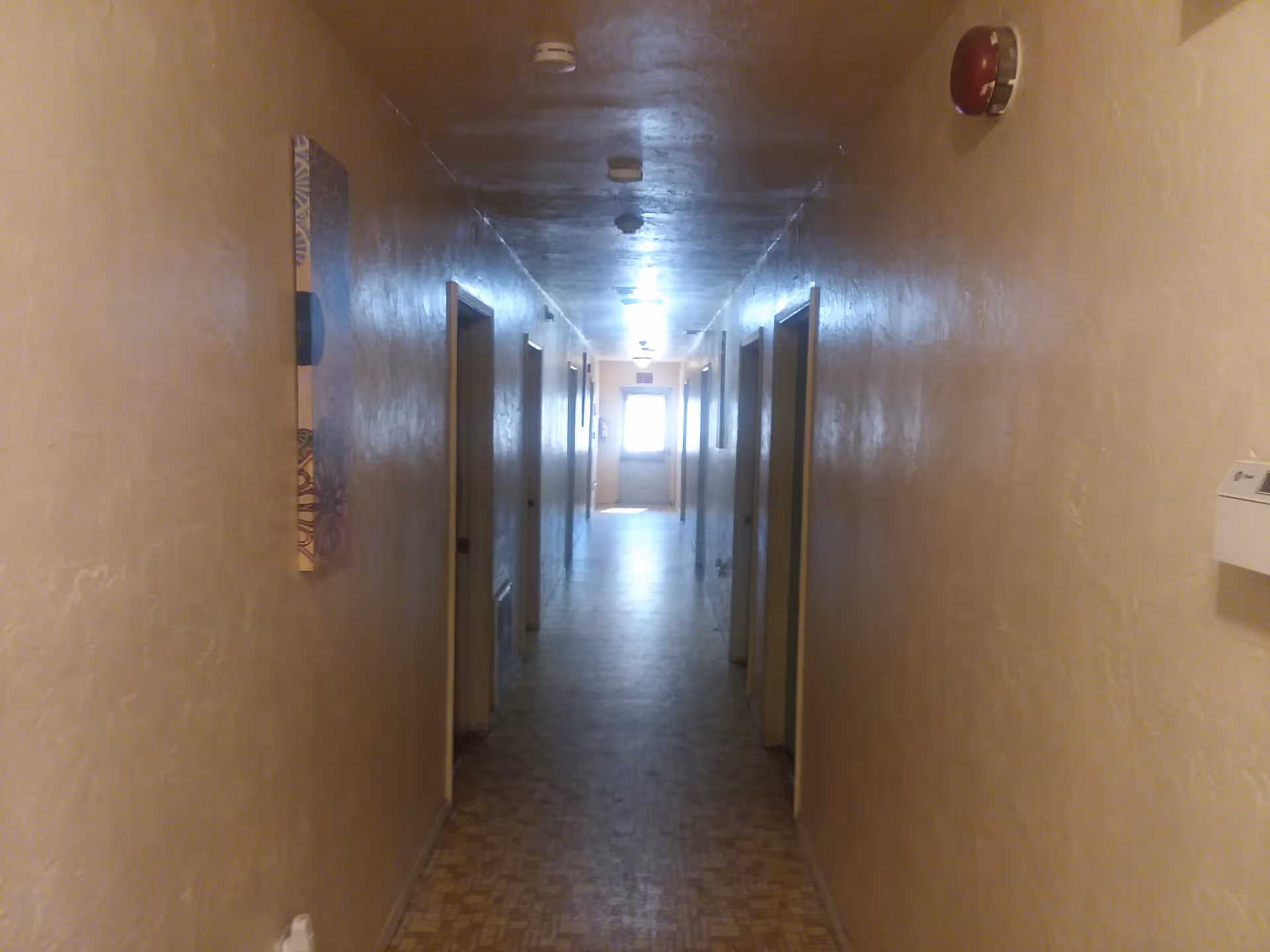 A long, narrow hallway with beige textured walls and a patterned floor. Several doorways line both sides of the hallway, and a bright window is visible at the far end. The ceiling has multiple light fixtures and smoke detectors.