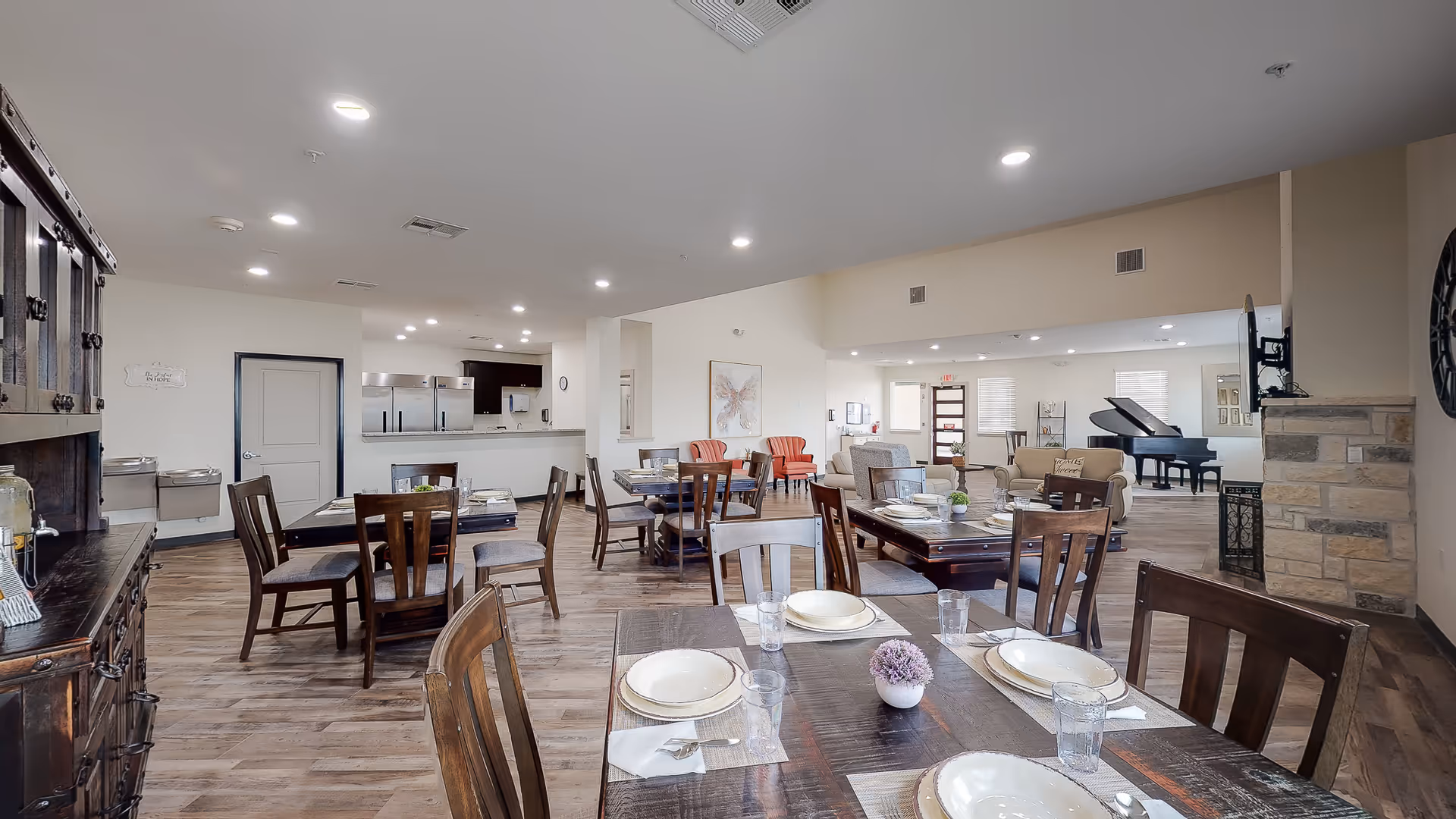 A spacious dining area in an assisted living facility with several wooden tables and chairs set with plates, glasses, and napkins. The room has wood flooring, bright overhead lighting, and a cozy seating area with armchairs and a grand piano in the background. There is a kitchen area visible with stainless steel appliances and a stone fireplace on the right side.