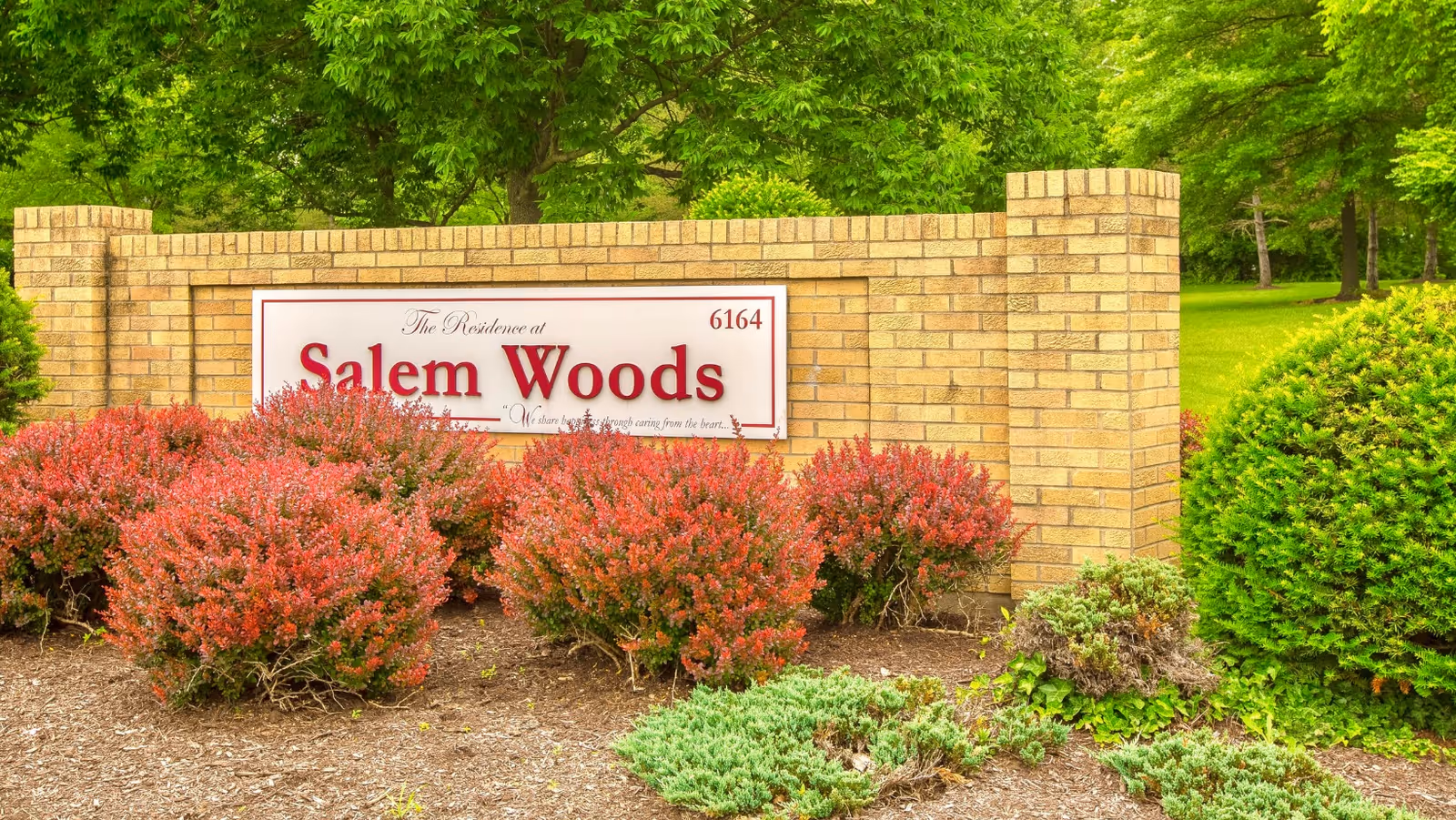 A brick sign for The Residence at Salem Woods surrounded by green trees and red and green shrubs in a landscaped garden area.