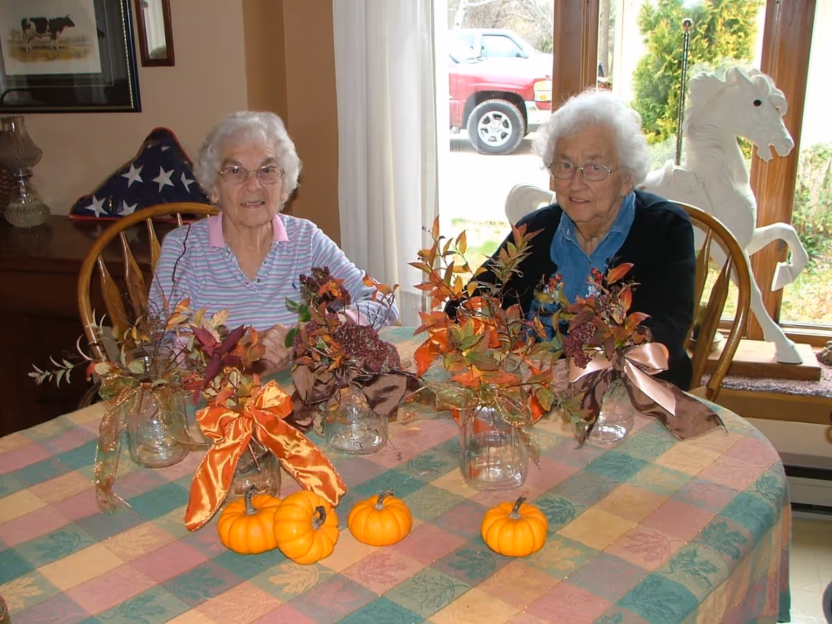 Two elderly women sit at a table decorated with small pumpkins and autumn floral arrangements in a dining area.
