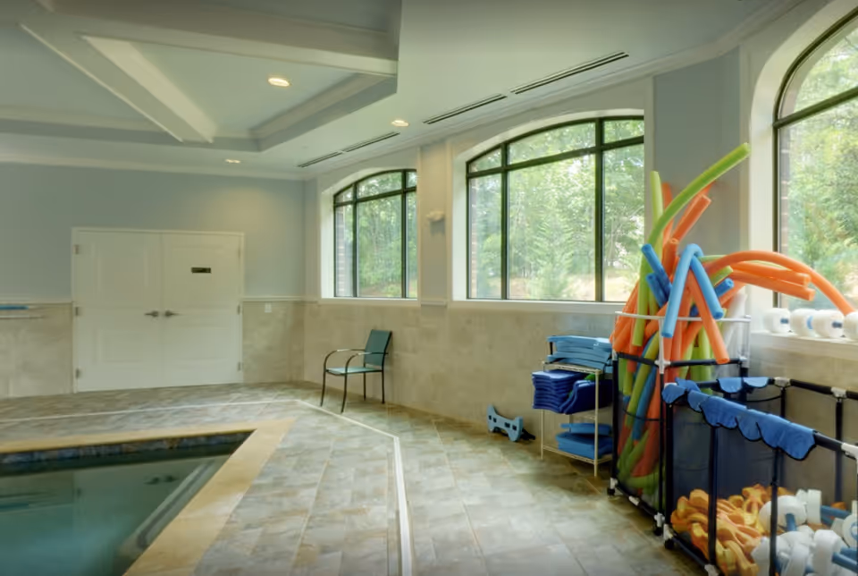 Indoor pool area with tiled floor and walls, large arched windows letting in natural light, a single chair against the wall, and racks holding colorful pool noodles and flotation devices.