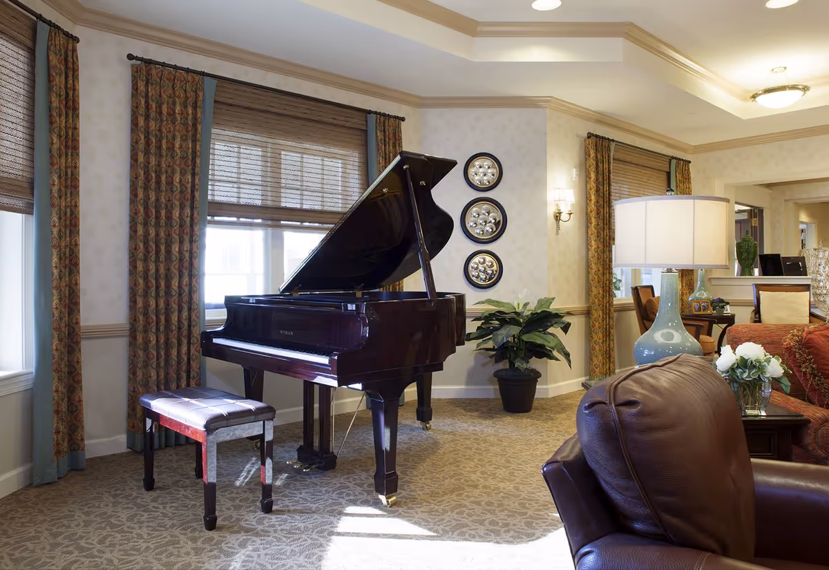 A cozy living room area featuring a grand piano with a matching bench near windows with patterned curtains. The room has beige patterned carpet, a leather armchair, a side table with a blue lamp, and a vase with white flowers. Decorative plates are mounted on the wall, and a potted plant is placed in the corner.
