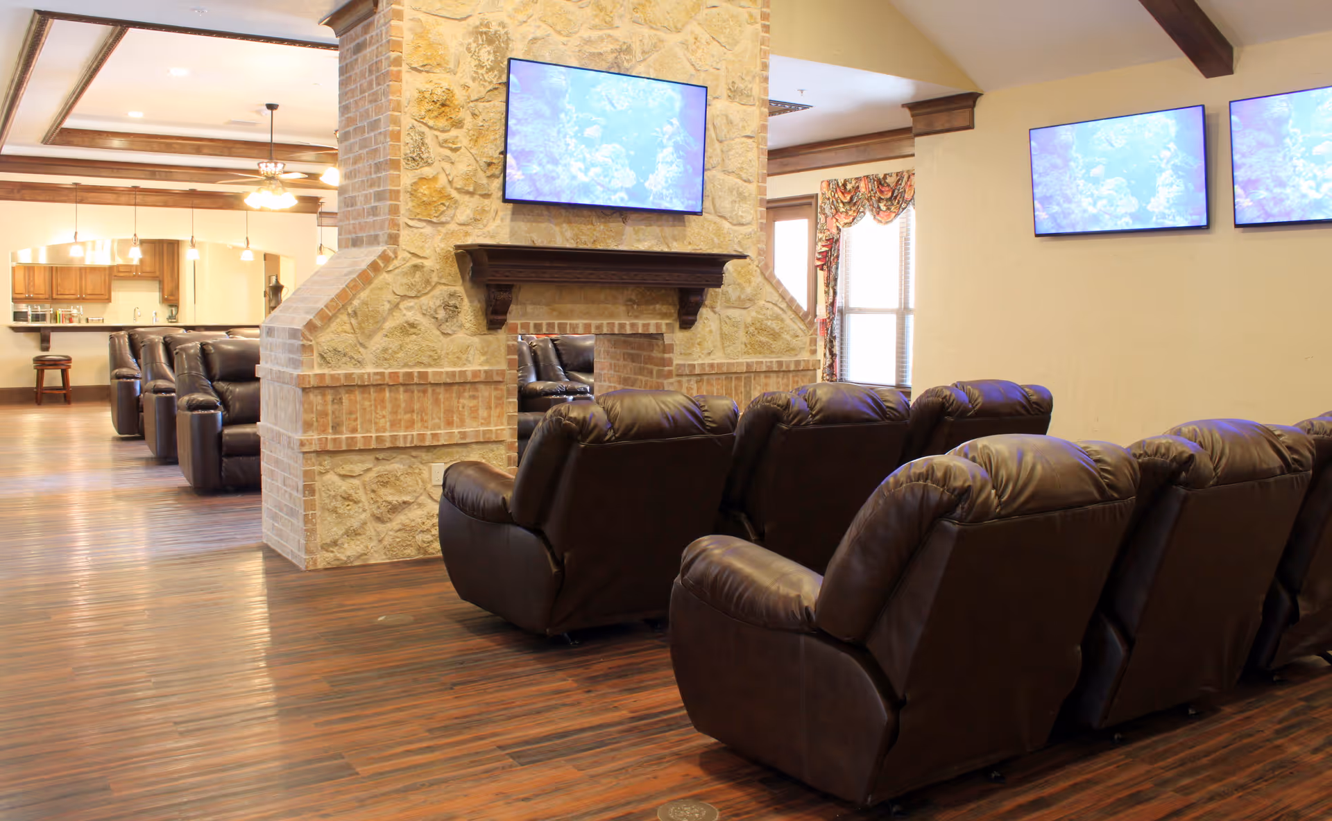 A memory-care common room with rows of brown leather recliners facing wall-mounted TVs above a stone fireplace and an open kitchen area in the background.
