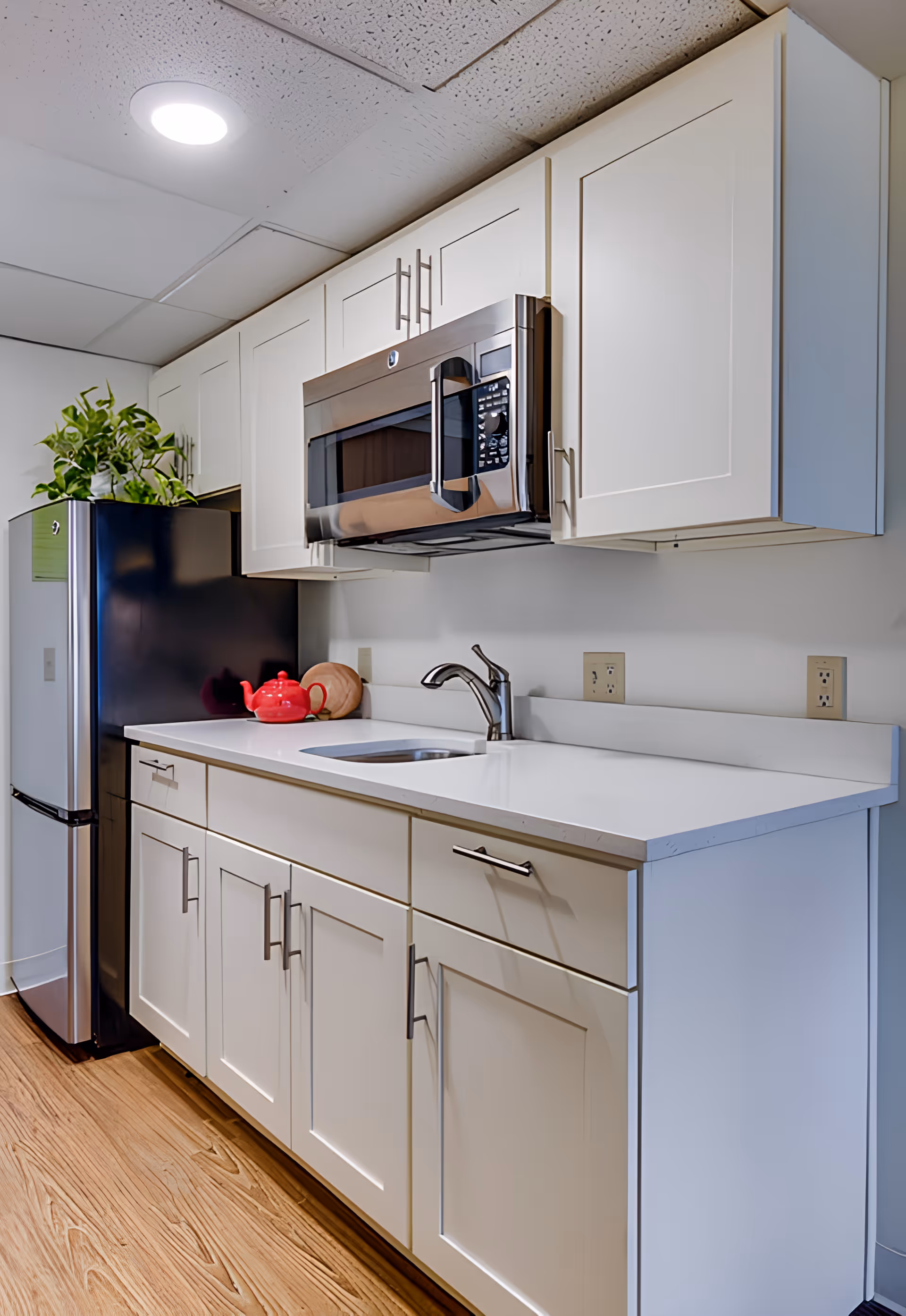 A modern kitchen area with white cabinets, a stainless steel microwave mounted above a white countertop with a sink and faucet. There is a stainless steel refrigerator with a green leafy plant on top. A red teapot and a wooden cutting board are placed on the countertop.