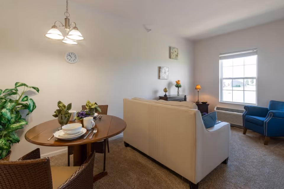 A cozy living and dining area with a round wooden dining table set for two, a beige sofa, a blue armchair near a window, and decorative plants and flowers. The room has beige carpeting and neutral-colored walls with a clock and wall art.