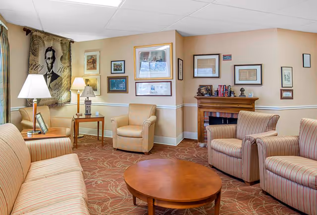 A cozy living room area with beige walls and carpet with a floral pattern. The room features a striped sofa, two striped armchairs, and a beige armchair arranged around a round wooden coffee table. There are two side tables with lamps, framed pictures and artwork on the walls, and a decorative fireplace with books and small sculptures on the mantel. A tapestry with an image of Abraham Lincoln hangs on one wall.