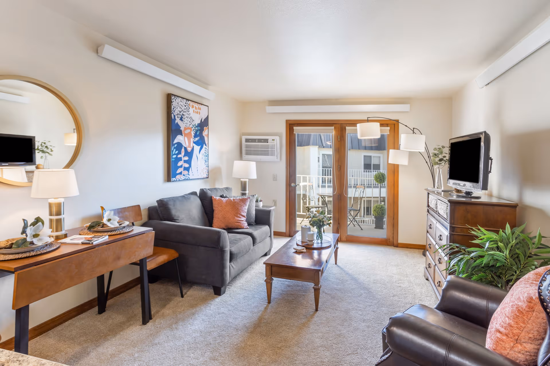 A cozy living room with a gray sofa, a wooden coffee table, a leather armchair with a pink cushion, a wooden dresser with a TV on top, and a small table with a lamp and decorative items. There is a large round mirror on the wall and a colorful abstract painting above the sofa. French doors lead to a balcony with outdoor furniture and potted plants.