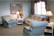 A cozy living room with two upholstered armchairs and a floral patterned sofa arranged around two white side tables with lamps. A window with floral curtains lets in natural light, and a framed artwork hangs on the wall above a side table with a flower arrangement.