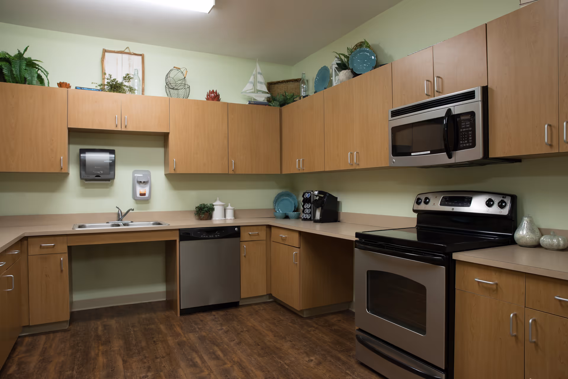 A clean and modern kitchen with wooden cabinets, stainless steel appliances including a dishwasher, oven, and microwave. The countertops are beige, and there are decorative items such as plants, plates, and a small sailboat model on top of the cabinets. The floor is a dark wood finish.