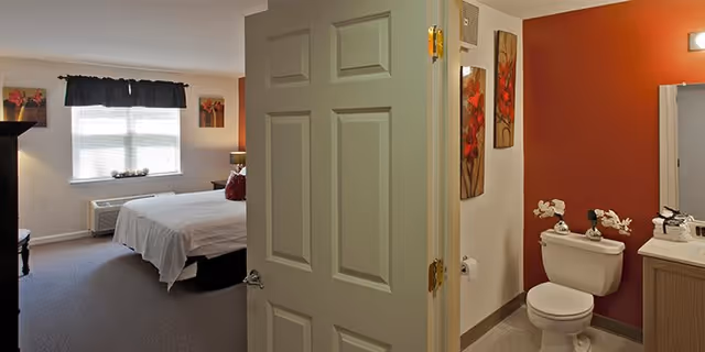 View of a senior living facility room showing a bedroom with a bed, window with curtains, and a small table, adjacent to a bathroom with a toilet, sink, and red accent wall.