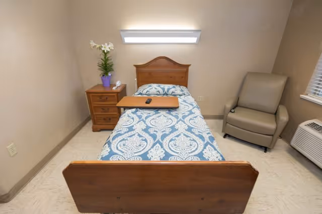 A small, simple bedroom with a single wooden bed covered with a blue and white patterned bedspread. There is a wooden nightstand with a purple flower pot and a telephone on it to the left of the bed. A beige armchair is positioned near a window with blinds on the right side of the room. An air conditioning unit is below the window. The room has beige walls and a tiled floor.