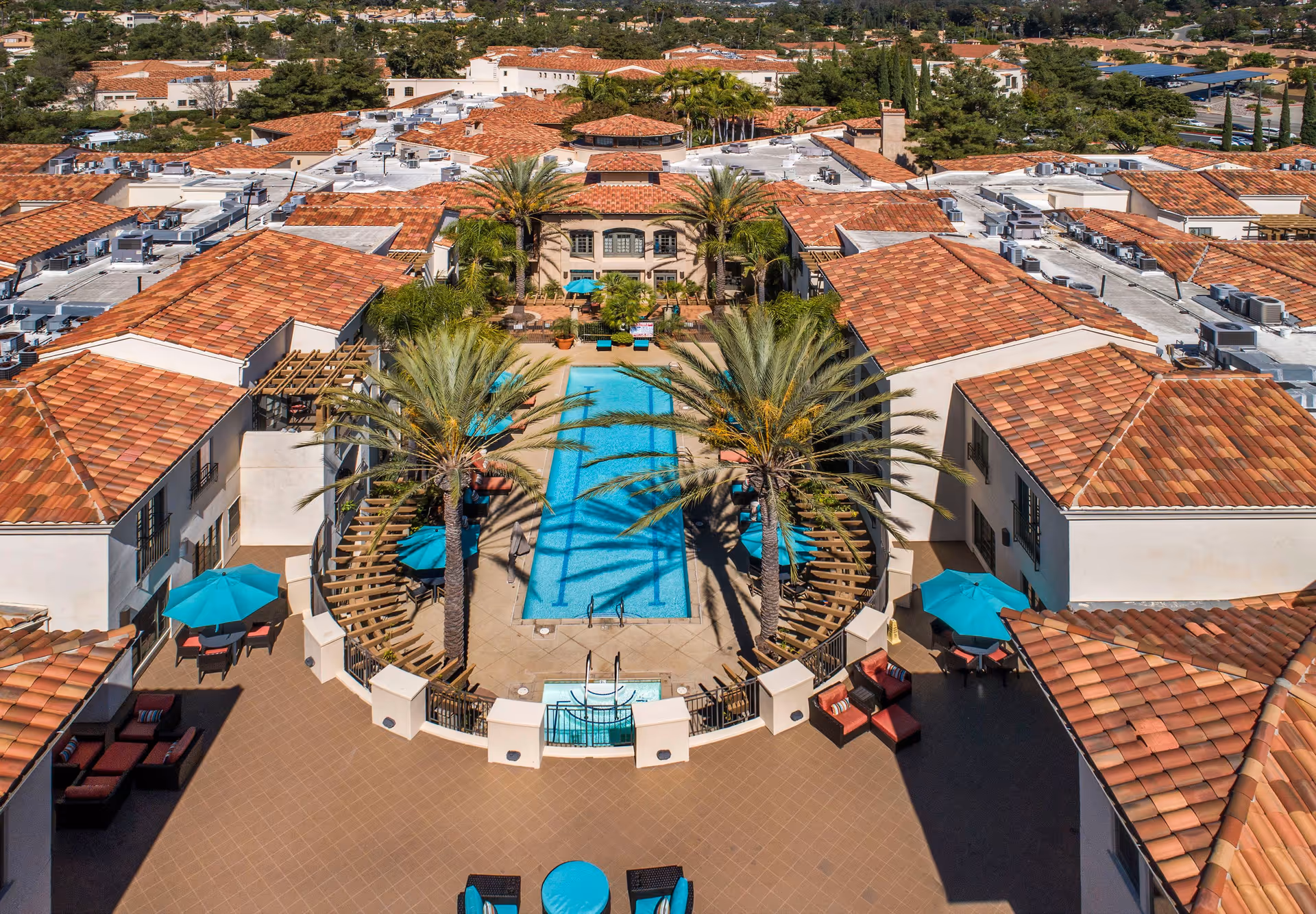 Aerial view of a senior living courtyard centered on a rectangular swimming pool with palm trees, turquoise umbrellas, and surrounding terracotta-roofed buildings.