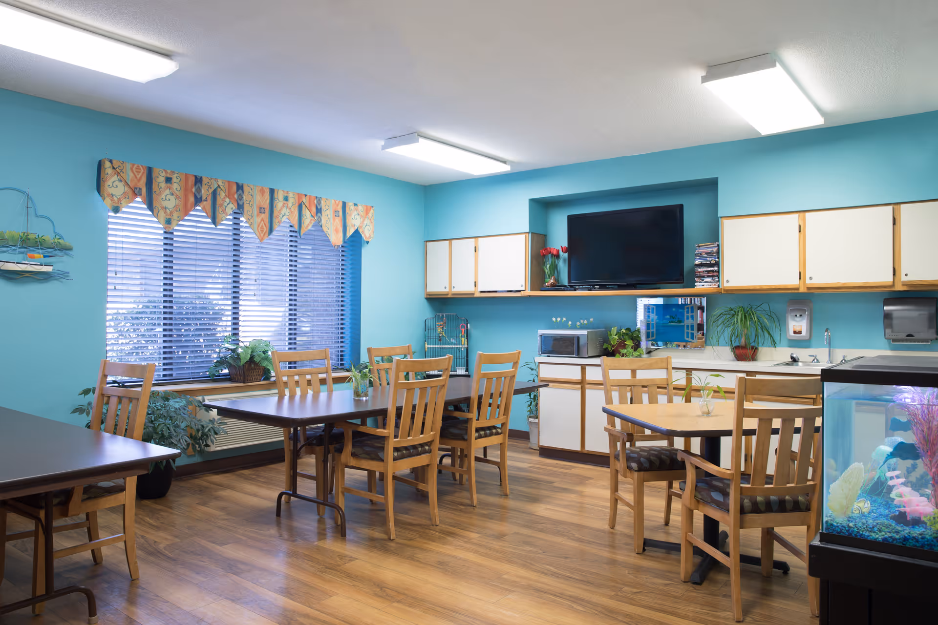 Bright communal dining/activity room with wooden tables and chairs, a kitchenette, wall-mounted TV, and an aquarium.