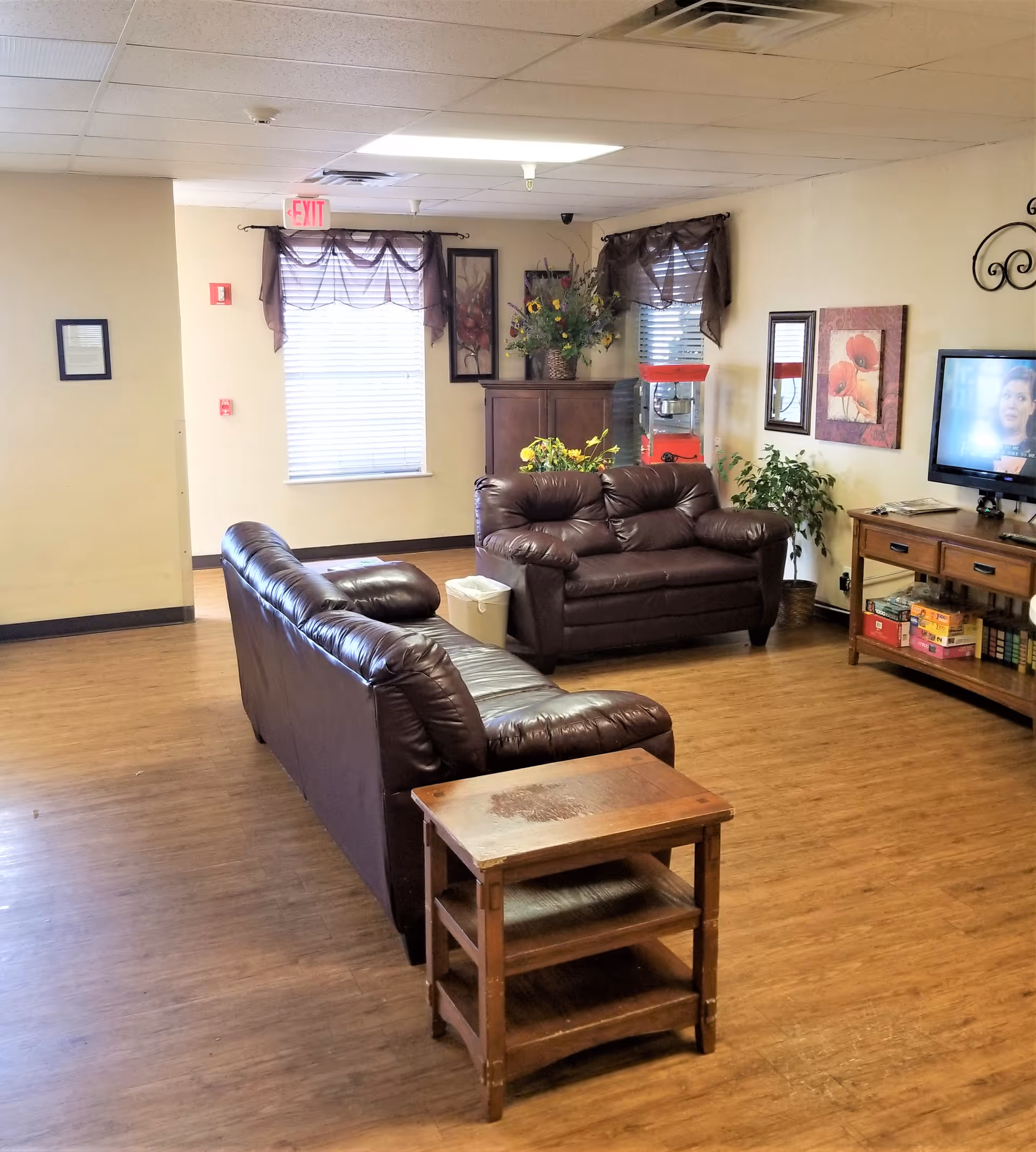 A cozy living room area in Georgia Manor Nursing Home featuring two brown leather sofas arranged around a wooden side table. The room has wood flooring, two windows with brown curtains, a wooden cabinet with a flower arrangement, a popcorn machine, a potted plant, and a TV mounted on the wall displaying a woman. The walls are decorated with framed artwork and a mirror.