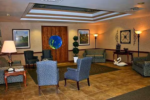 Lounge area with upholstered chairs, lamps, framed art, and a wooden door in a senior facility.