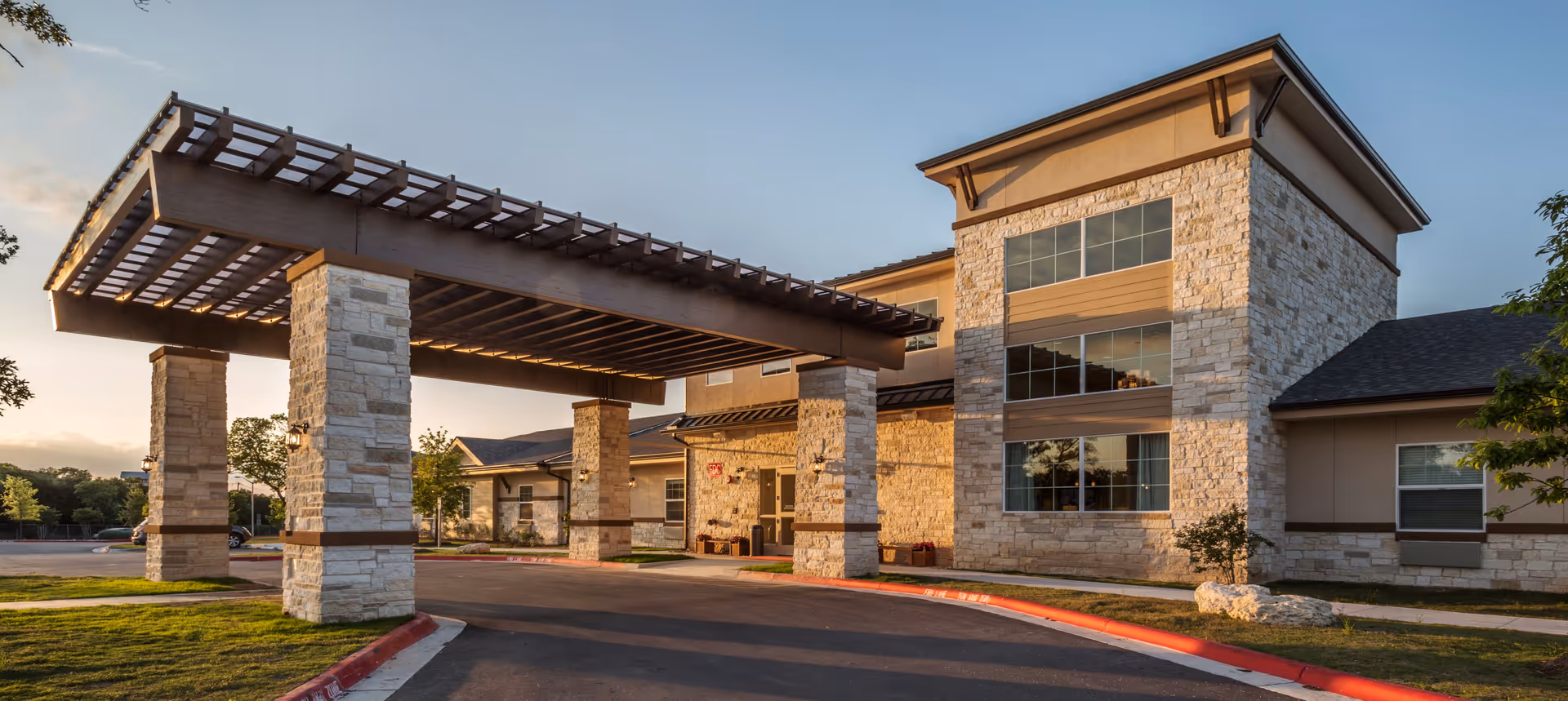 Exterior view of a senior living facility building with stone pillars and a large covered entrance area under a wooden pergola structure, surrounded by a driveway and landscaped grass areas during sunset.