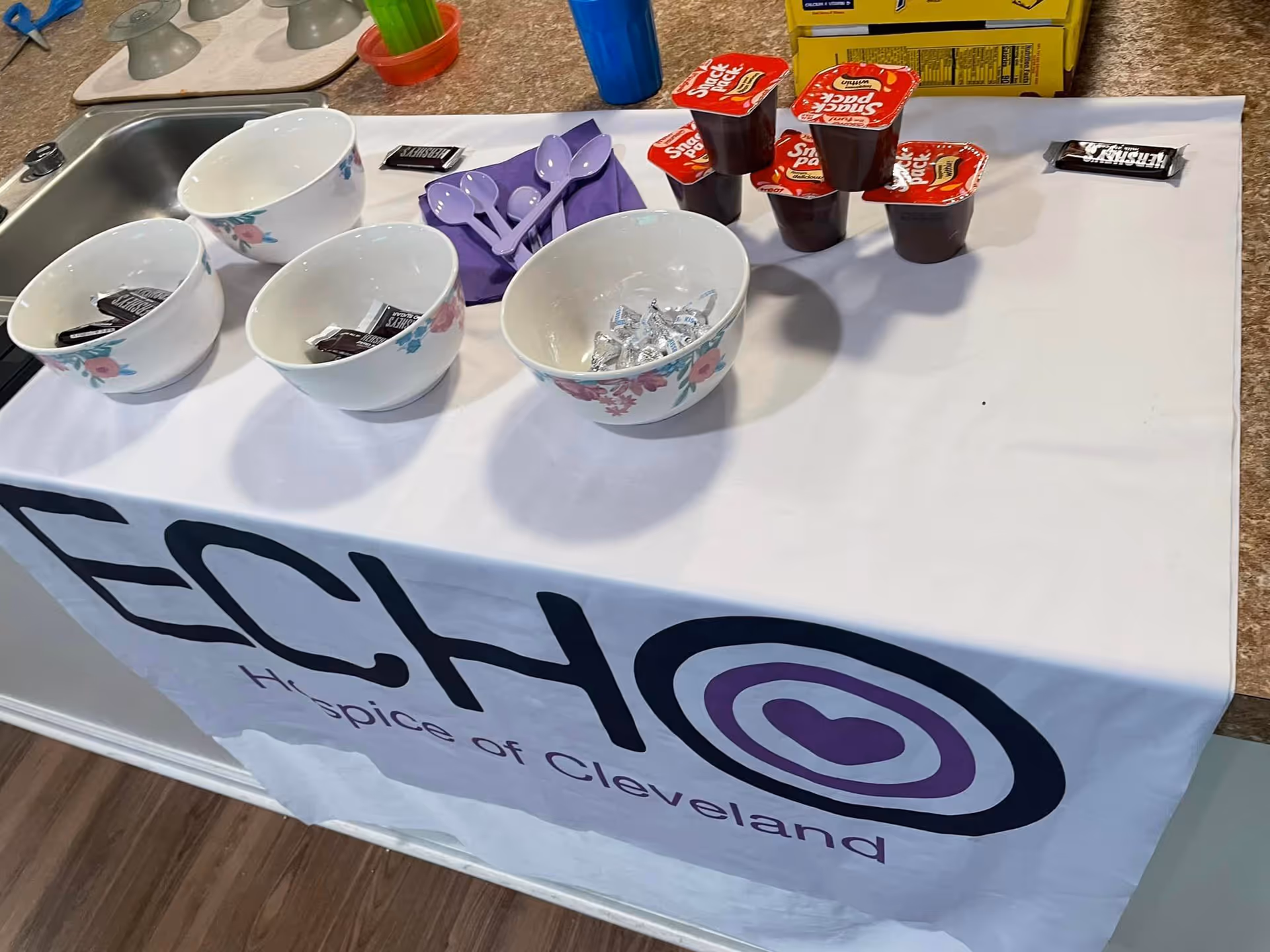 A countertop with a white tablecloth displaying the logo and text 'ECHO Hospice of Cleveland'. On the counter are five white bowls with floral designs, some containing Hershey's chocolate bars and Hershey's Kisses. There are also several Snack Pack pudding cups stacked in a pyramid, purple plastic spoons on a purple napkin, a blue cup, and a yellow box in the background.