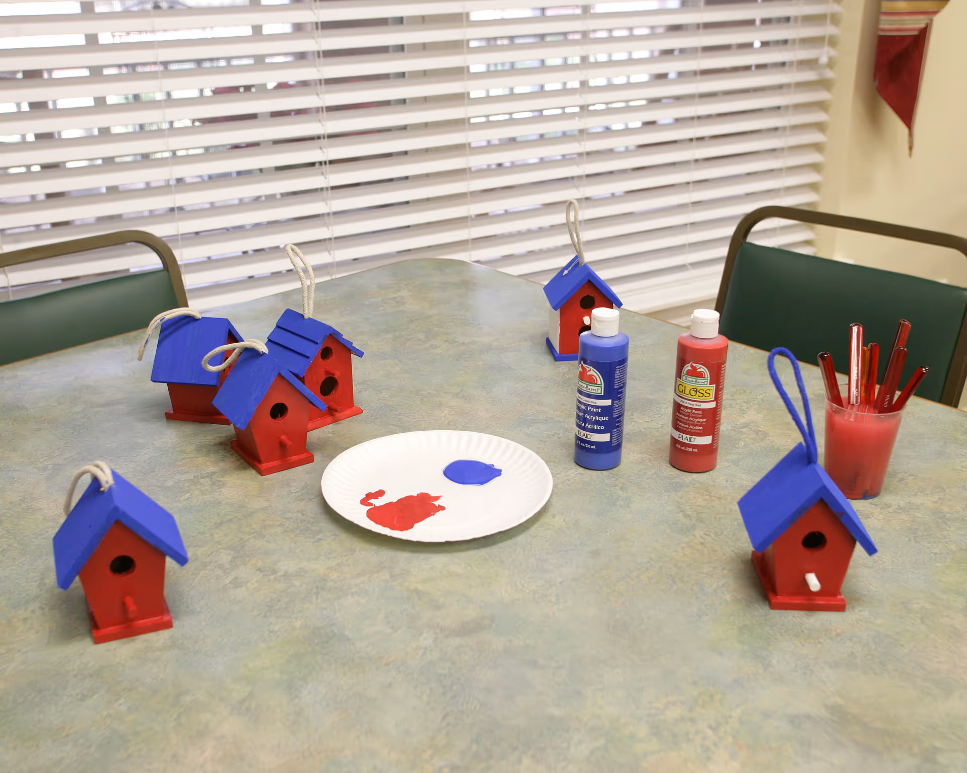 A table with several small red and blue painted birdhouses, two bottles of paint (one blue and one red), a paper plate with red and blue paint, and a cup holding red paintbrushes. The background shows window blinds and green chairs around the table.