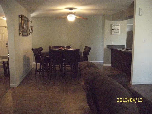Interior view of a living and dining area in a senior living facility. The image shows a dining table with six chairs under a ceiling fan with a light. To the right, there is a partial view of a kitchen counter and a brown couch in the foreground. The walls are light-colored, and there is a decorative wall hanging and a framed sign on the walls.