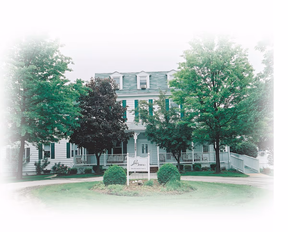 Front exterior view of LifeSpring Assisted Living facility, a white multi-story building with green shutters, surrounded by trees and greenery, with a sign in front displaying the facility name.