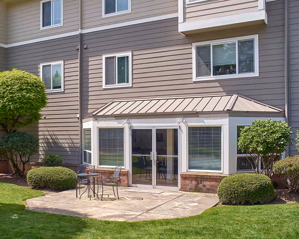 Exterior view of a senior living building's ground-floor patio with sliding glass doors, a small table and chairs, and trimmed shrubs.