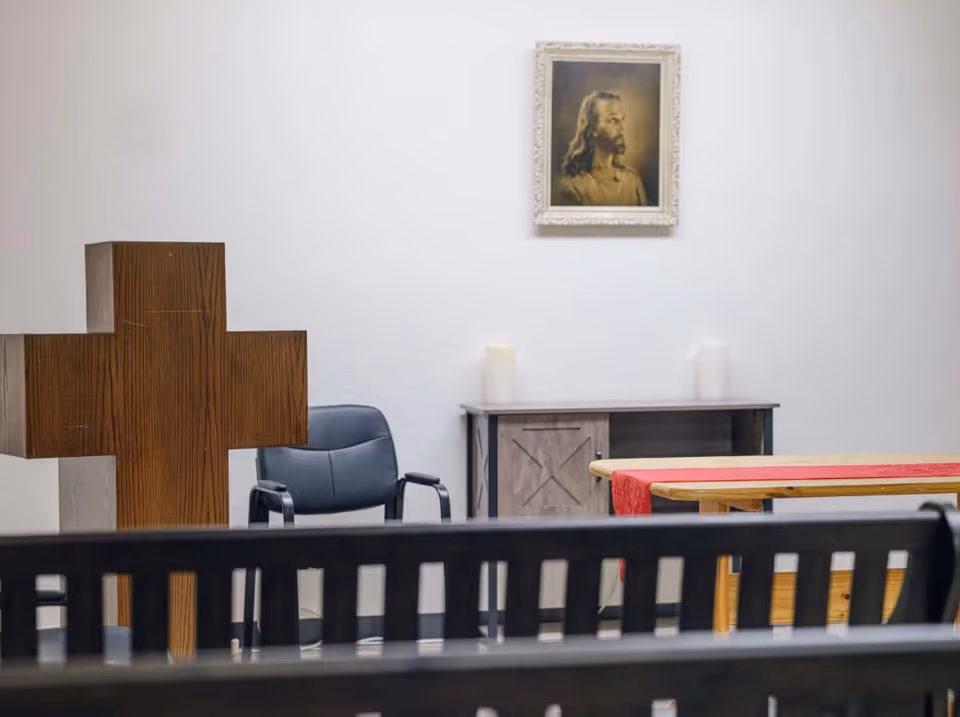 Interior view of a small chapel or prayer room with a large wooden cross, a black chair, a cabinet with two candles on top, a table covered with a red cloth, and a framed picture of Jesus on the white wall.