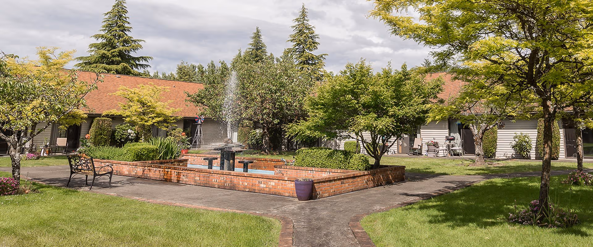 Outdoor courtyard area with a brick fountain in the center, surrounded by green grass, trees, and shrubs. There are benches along the paved pathways and single-story buildings with red roofs in the background under a cloudy sky.