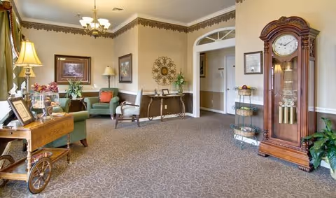 A spacious and elegantly decorated senior living facility common area with beige walls and carpeted floor. The room features a large wooden grandfather clock on the right, a small table with plants and framed pictures, a green armchair with a red cushion, and a wooden side table with a lamp and decorative items. There is a chandelier hanging from the ceiling and framed artwork on the walls.