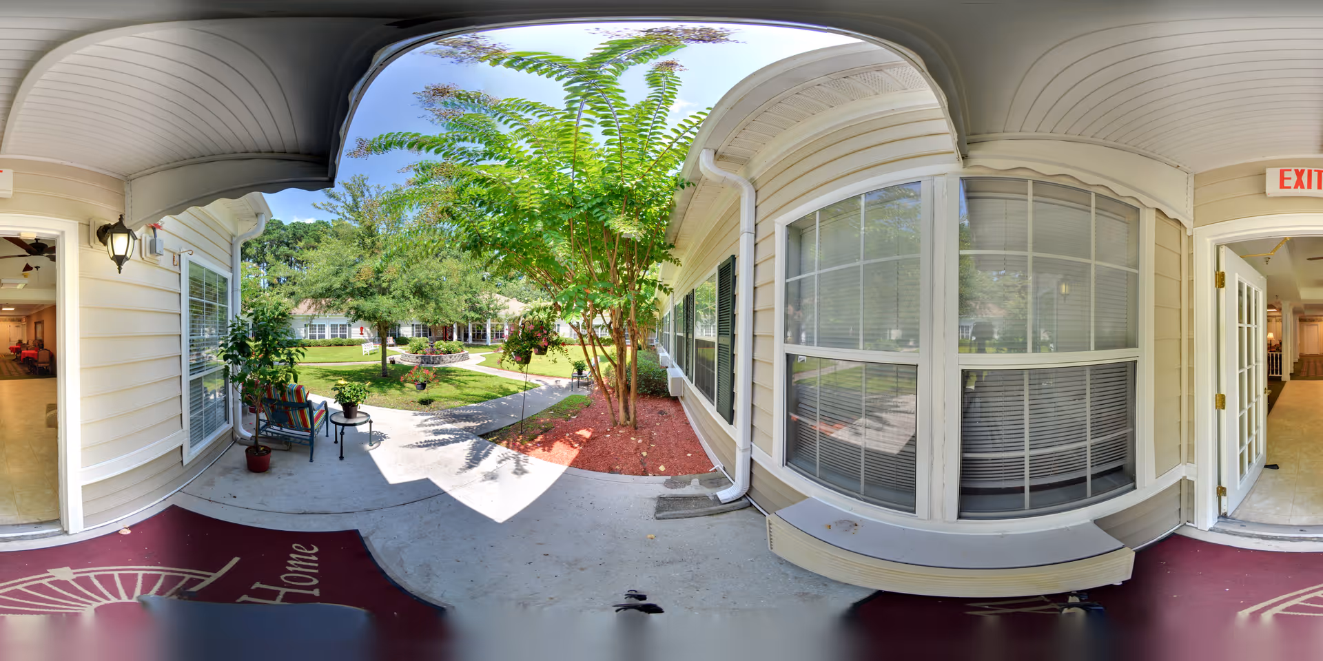 Covered porch area at the entrance of a senior living facility with a bench, potted plants, and a view of a landscaped garden with trees and a walking path. The porch has white siding, large windows, and an open door leading inside. The sky is clear and blue.