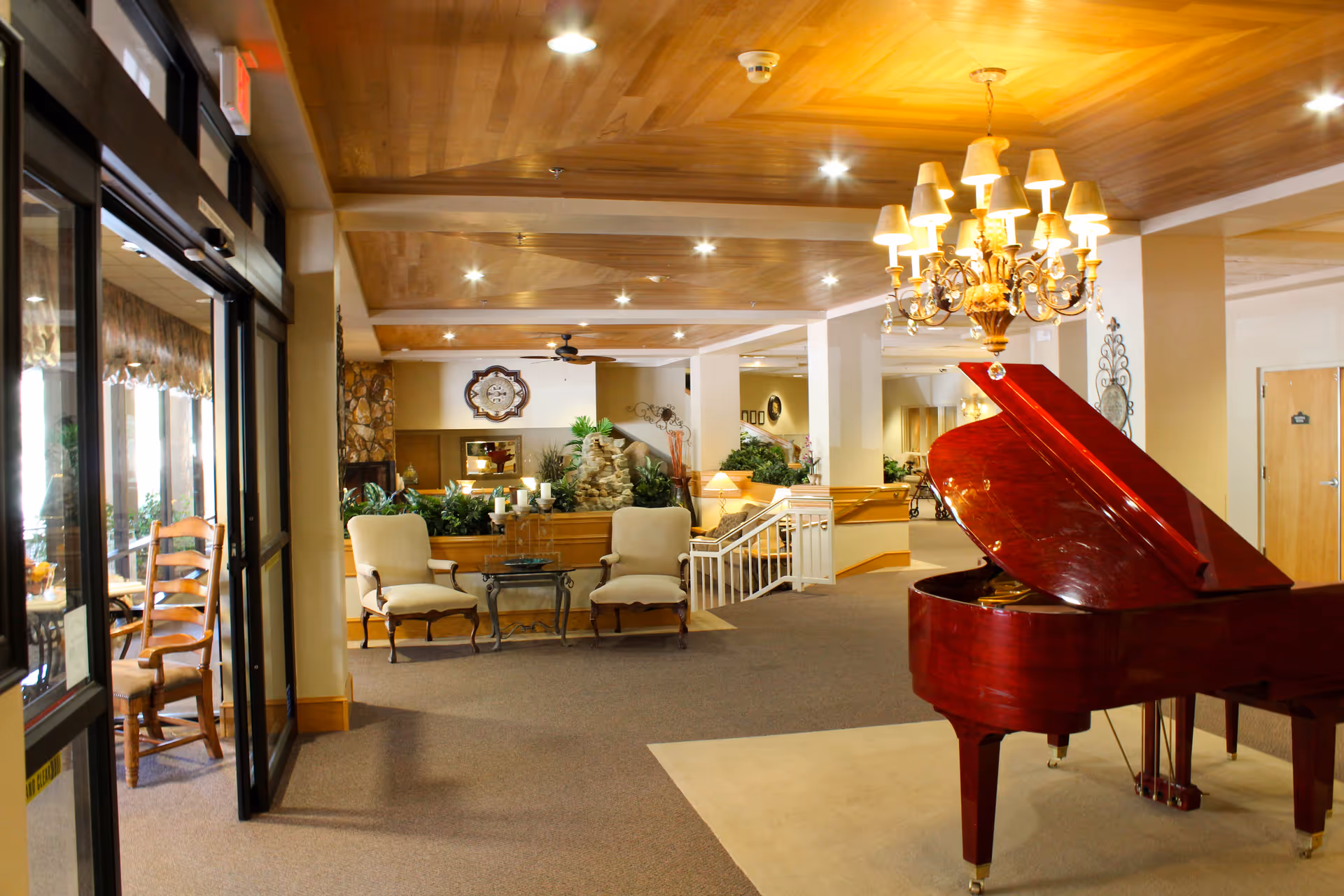 Lobby of a senior living facility featuring a red grand piano, chandelier, seating area, and glass entrance.
